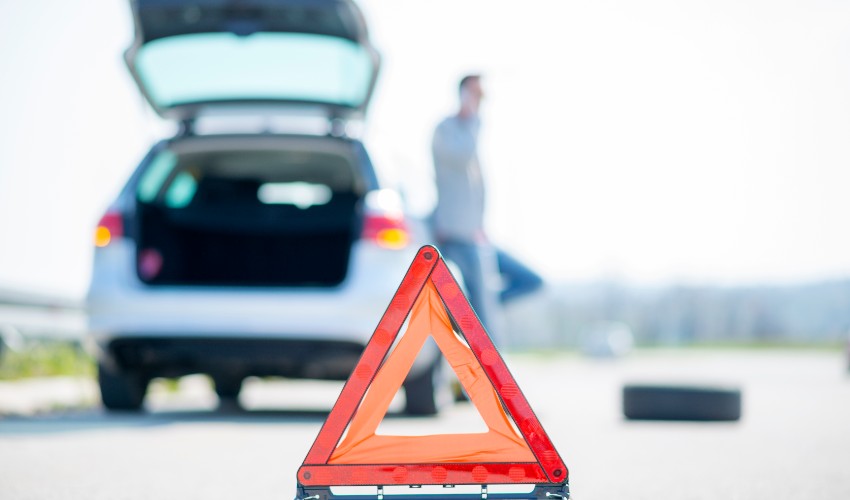 A man with a silver car that broke down on the side of the road. He put up a red warning triangle and is waiting for the technician to arrive.