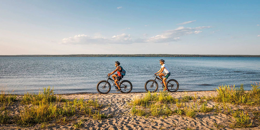 Two cyclists ride along a sandy beach trail dotted with grass, under a blue sky with clouds on a sunny day.