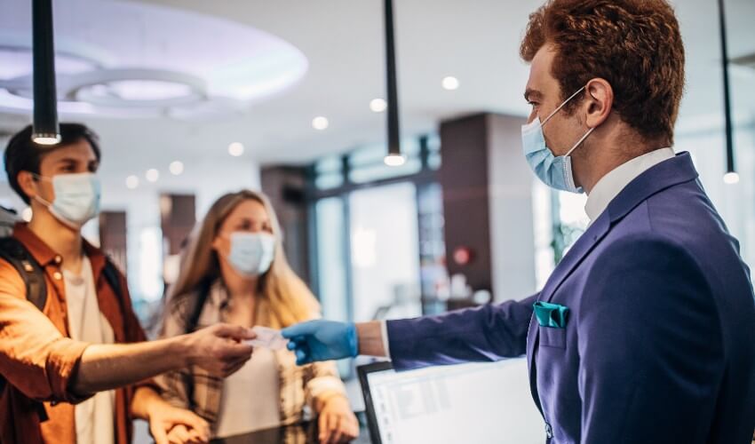 Couple wearing non-medical face masks speaking with a male hotel receptionist at front desk. 